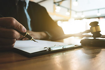 man in suit at a desk with a clipboard in front of him, pen in hand, and a gavel on his desk. 