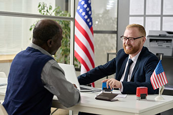 government official sitting across the desk from his constituent with a U.S. flag on his desk and behind him