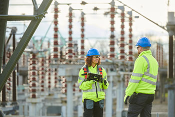 a woman and a man utility worker in high-viz jackets and hard hats standing in front of electrical grid.